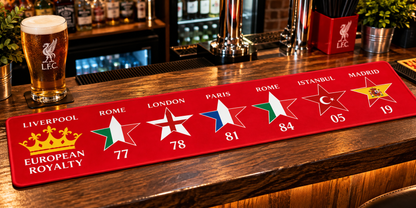 A red bar mat featuring the UEFA Champions League final host cities and years (Liverpool, Rome, London, Paris, Rome, Istanbul, Madrid) with club crests and gold stars, displayed on a wooden bar counter next to a glass of beer.