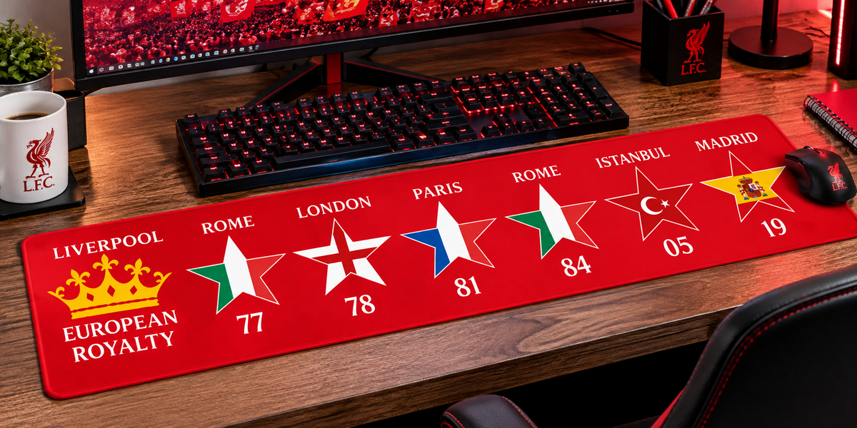 A large red gaming desk mat featuring multiple football club and city logos with star graphics and numbers, shown on a wooden desk in front of a keyboard and monitor.