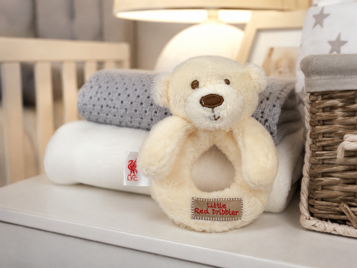 A cream-colored plush teddy bear ring rattle with a stitched 'Little Red Trumper' label, displayed on a white shelf next to a wicker basket and folded baby blankets.