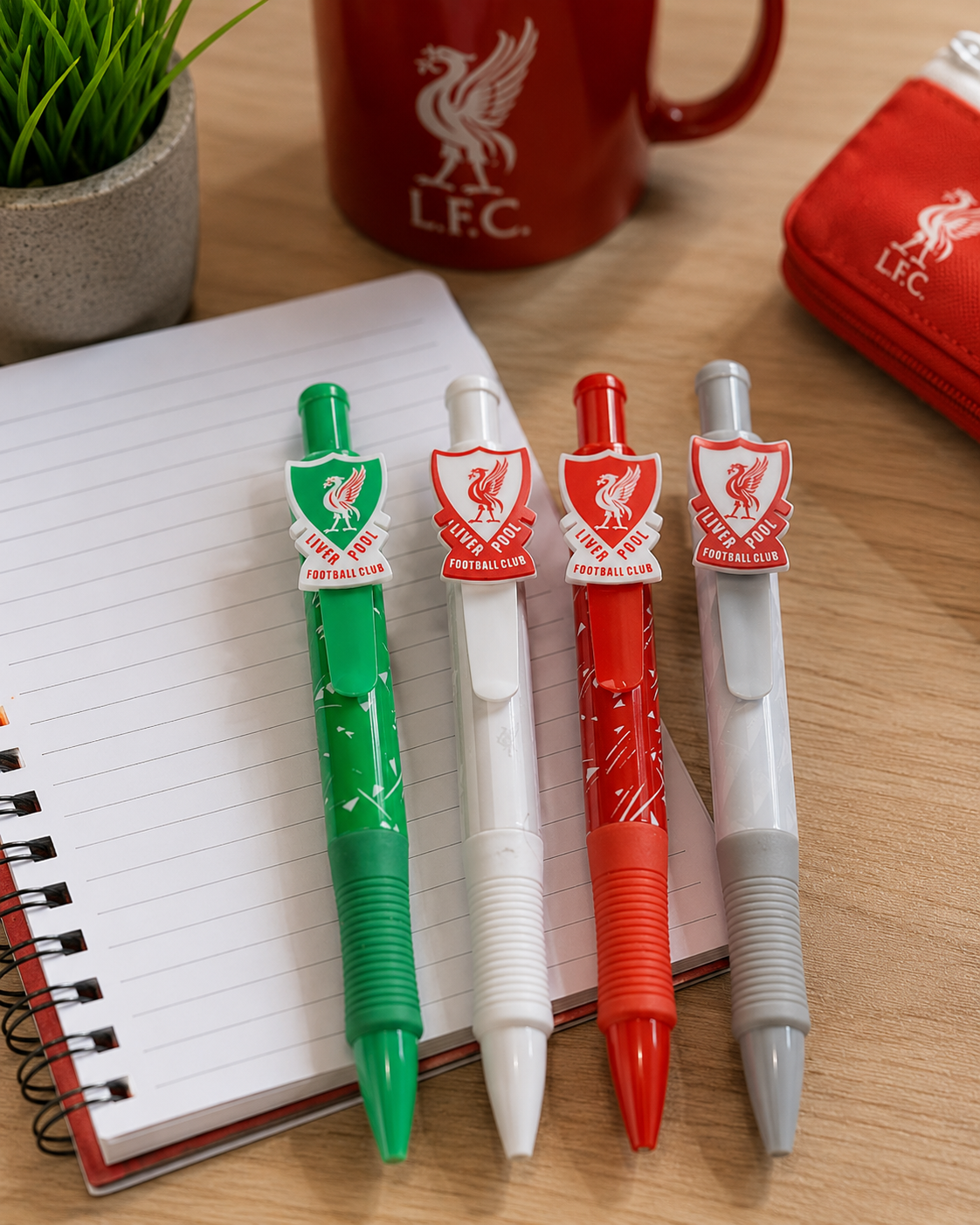 Four Liverpool Football Club themed retractable pen markers in green, white, red, and gray, lined up on an open notebook. A red L.