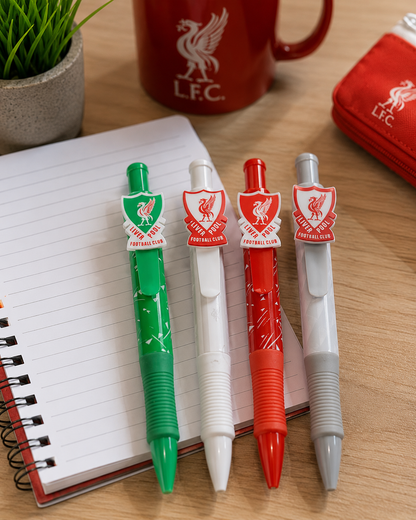Four Liverpool Football Club themed retractable pen markers in green, white, red, and gray, lined up on an open notebook. A red L.