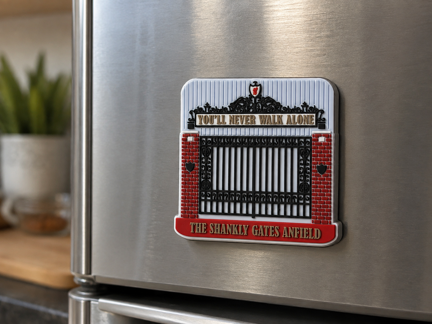 A rectangular refrigerator magnet featuring the Anfield gate design from 'You'll Never Walk Alone' by The Shanks, shown attached to a stainless steel fridge in a kitchen setting.